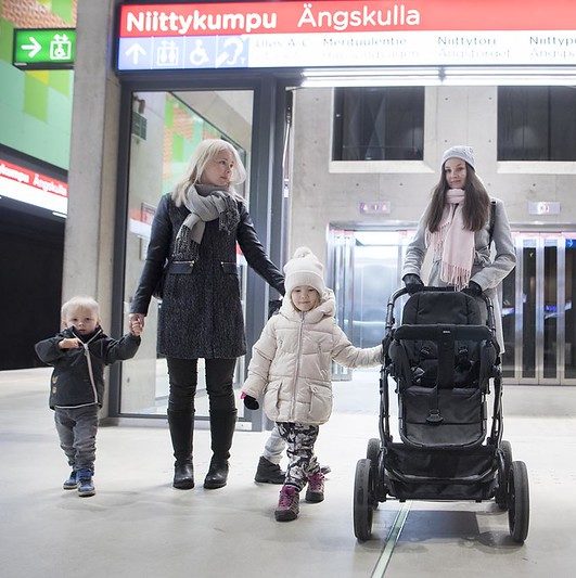 Kuvassa on kaksi naista ja kolme lasta metroasemalla lastenrattaiden kanssa. Taustalla näkyy metroaseman opasteet.